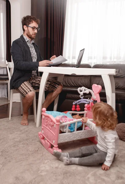 A young man Businessman making Video Conferencing at the Laptop from home.  Wearing Strong jacket, shirt and home shorts. Little daughter sitting near the father and plays dolls.