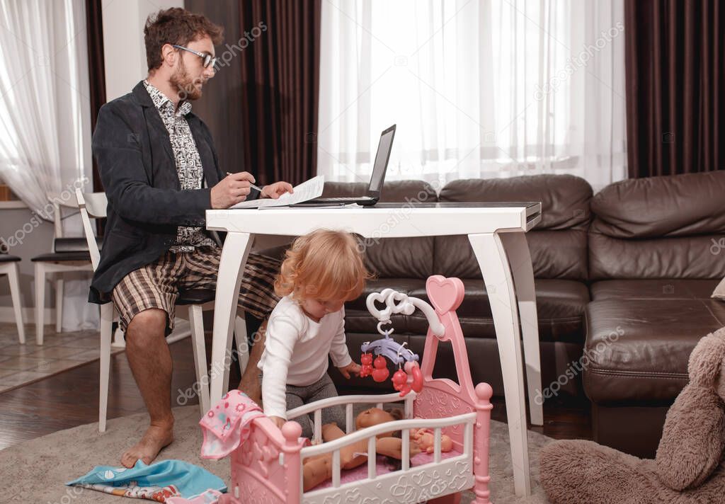A young man Businessman making Video Conferencing at the Laptop from home.  Wearing Strong jacket, shirt and home shorts. Little daughter sitting near the father and plays dolls.