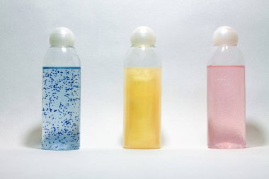 Üç cam duş jeli şişesi beyaz kapaklı, farklı renklerde ve açık arkaplanda ışıltılar var. Mavi, altın ve pembe renkteler.. 