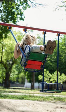 Sarışın kız güneşli bir havada baharda parkta sallanıyor. Kot pantolon ve beyaz bluz giyiyor.