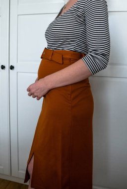 Evde turuncu etekli ve göbeğini tutan çekici hamile bir kadın. Annelik, hamilelik, mutluluk..