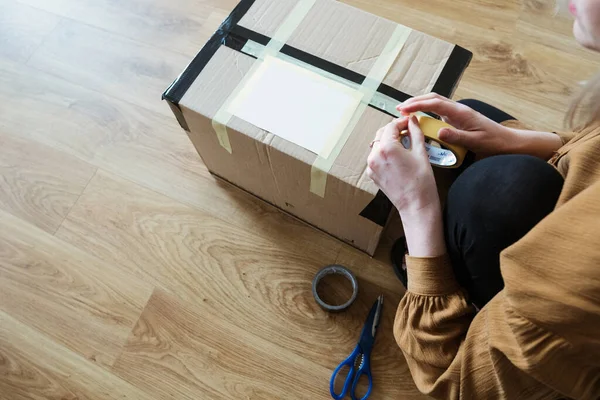 Müşteri olan genç bir kadın paket hazırlıyor. Bant ve makas kullanarak. Kız tüketici yerde karton kutu tutuyor..