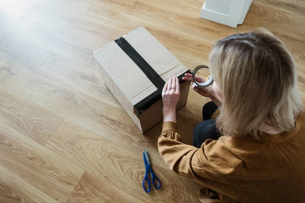 Müşteri olan genç bir kadın paket hazırlıyor. Bant ve makas kullanarak. Kız tüketici yerde karton kutu tutuyor..