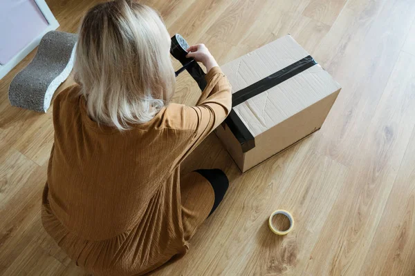 Müşteri olan genç bir kadın paket hazırlıyor. Bant ve makas kullanarak. Kız tüketici yerde karton kutu tutuyor..