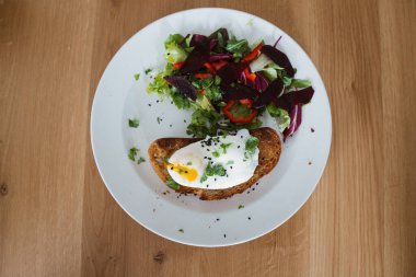 Bir parça ekmeğin üzerinde haşlanmış yumurta, tabakta yeşil salata ve ahşap masanın üzerinde. Sağlıklı Kahvaltı