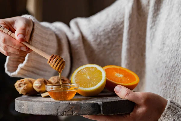 Young woman in holding immunity strengthening kit: ginger, lemon, orange, honey pourring from spoon. Anti virus natural organic treatment. Healthcare, home remedy against cold, influenza. Closeup