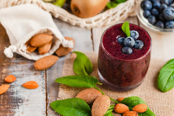 Healthy food and vegan diet concept - glass of fresh juice or smoothie with blueberry, spinach, banana, almond milk. Antioxidant detox beverage with raw ingredients.  Close up, wooden background.
