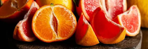 Slices of ripe fresh organic citrus fruits: grapefruit, orange, lemon on wooden board. Natural source of vitamins, low calories tasty dessert. Dark background, close up, front view, macro. Banner