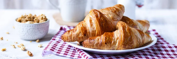 Delicious breakfast french style at home: light background, white plate with fresh croissants, small bowl of granola, cup of tea and bottle with milk. Served on red and beige napkins. Banner