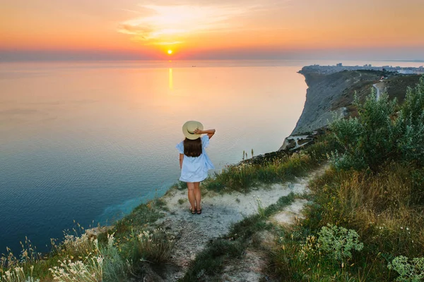 Mavi elbiseli bir kız uçurumun kenarında duruyor ve gün batımını seyrediyor.
