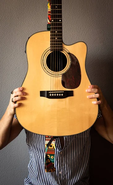Renkli gömlekli ve klasik yüzüklü genç bir adam yüzünü bir gitarla örtüyor. Gri arka plan. Seçici odak.
