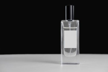 Siyah arka planda beyaz bir masanın üzerinde bir şişe parfüm duruyor. Metniniz için boş bir etiket var