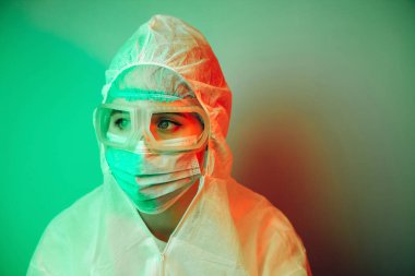 Siyah arka planda koruyucu giysili genç kız doktor. Kasvetli ışıklandırma. Yorgun doktor. Salgın covid-2019. Gözlüklü, eldivenli ve maskeli bir doktorun portresi. Koronavirüsün yayılmasını engelle.
