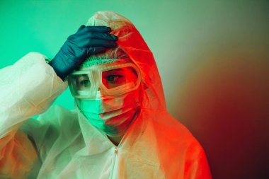 Siyah arka planda koruyucu giysili genç kız doktor. Kasvetli ışıklandırma. Yorgun doktor. Salgın covid-2019. Gözlüklü, eldivenli ve maskeli bir doktorun portresi. Koronavirüsün yayılmasını engelle.