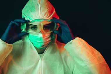 Siyah arka planda koruyucu giysili genç kız doktor. Kasvetli ışıklandırma. Yorgun doktor. Salgın covid-2019. Gözlüklü, eldivenli ve maskeli bir doktorun portresi. Koronavirüsün yayılmasını engelle.