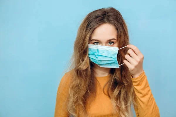 Sarı tişörtlü bir kız tıbbi maske takıyor. Mavi arka planda, koruyucu maskeli bir sarışının portresi. Koronavirüsten koruma. Covid-2019. salgın 2020.