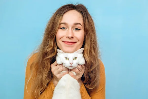 Genç bir kız kollarında beyaz bir kedi tutuyor. Mavi arka planda kıvırcık saçlı sarışın bir kızın portresi. Hayvan koruma kavramı. Sığınaktan kediyi al..