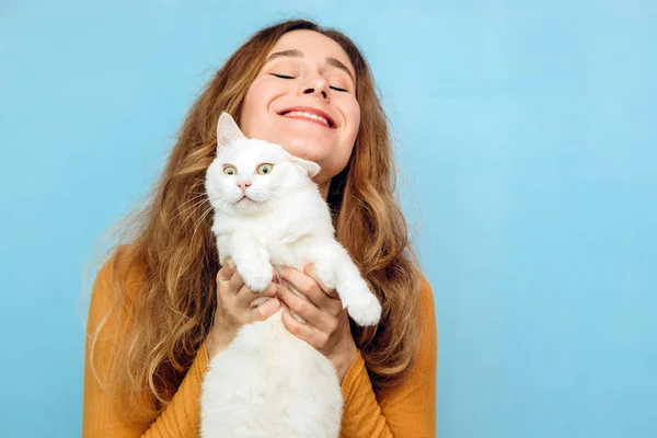 Genç bir kız kollarında beyaz bir kedi tutuyor. Mavi arka planda kıvırcık saçlı sarışın bir kızın portresi. Hayvan koruma kavramı. Sığınaktan kediyi al..