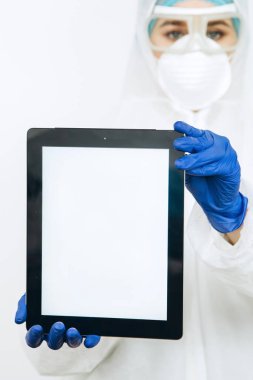 Koruyucu giysili doktor, solunum cihazı, gözlük, eldiven bir tablet / telefon tutuyor. Mesaj için yer. Doktorlar koronavirüs salgını sırasında evde kalmalarını tavsiye ettiler. COVID-19