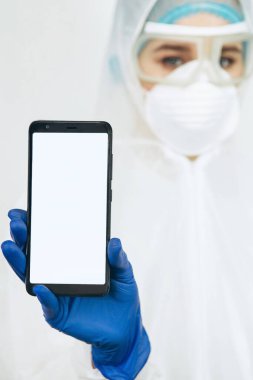 Koruyucu giysili doktor, solunum cihazı, gözlük, eldiven bir tablet / telefon tutuyor. Mesaj için yer. Doktorlar koronavirüs salgını sırasında evde kalmalarını tavsiye ettiler. COVID-19
