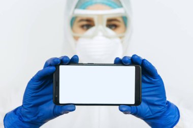Koruyucu giysili doktor, solunum cihazı, gözlük, eldiven bir tablet / telefon tutuyor. Mesaj için yer. Doktorlar koronavirüs salgını sırasında evde kalmalarını tavsiye ettiler. COVID-19