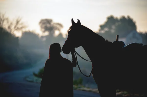一个穿着雨衣的女孩在乌云笼罩的背景下亲吻一匹马的轮廓 骑手和马在黎明时分 — 图库照片