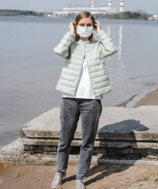 Açık renk ceketli bir kız güneşli bir günde nehrin arka planında maske takıyor.