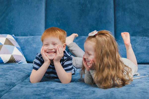 A little girl with red hair looks at a boy with red hair who is smiling. Brother and sister are lying on a blue sofa.