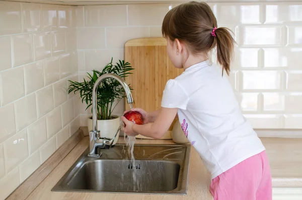 Girl washes a red apple in the kitchen near the sink. The concept of childrens health and independence