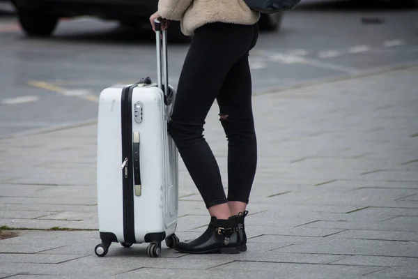 Девушка в черных джинсах и черных туфлях ждет на улице с белым чемоданом — стоковое фото