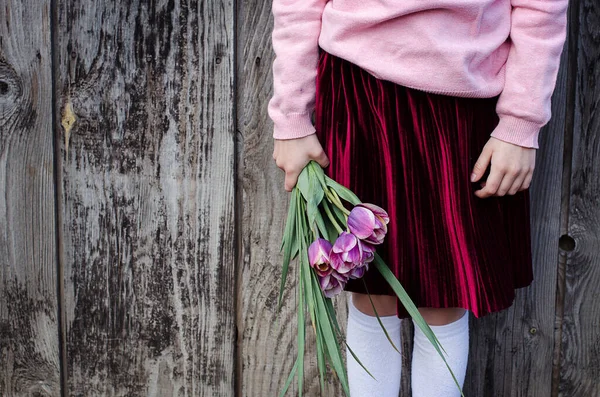 A girl in white knee socks and burgundy pleated skirt holds in her hand a bouquet of purple tulips. Old wood background. Copy space. Concept of spring bouquet. Front view.