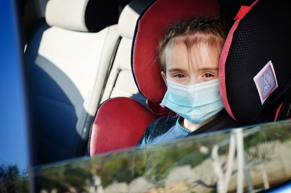 Tıbbi maskeli küçük bir kız arabada çocuk koltuğunda oturuyor. Covid-19 Coronavirus pandemik küresel yayılma konsepti. Maskeli bir çocuğun portresi.
