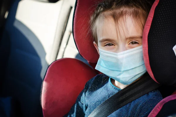 Tıbbi maskeli küçük bir kız arabada çocuk koltuğunda oturuyor. Covid-19 Coronavirus pandemik küresel yayılma konsepti. Maskeli bir çocuğun portresi.