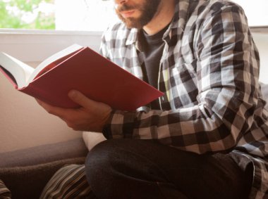 Karantina sırasında ekose gömlek giyen sakallı adam gün ışığında kanepede kırmızı bir kapak kitabı okurken pencereden içeri giriyor..