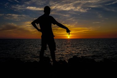 Ayakta duran bir adamın silueti gün batımından sonra parmağını deniz kenarındaki güneşe doğrultur..