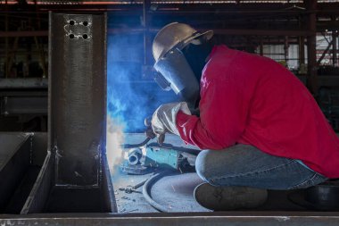 Kaynakçı, Flux Cored Arc Welding (FCAW) işlemiyle bir çelik yapı çalışması yapıyor ve endüstriyel fabrikada güvenlik için kişisel koruyucu ekipman (PPE) ile uygun şekilde giyiniyor.