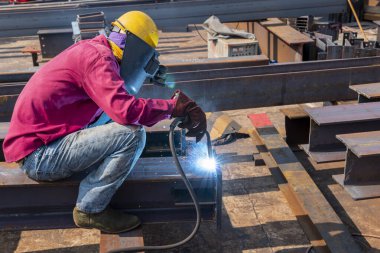 Kaynakçı, Flux Cored Arc Welding (FCAW) işlemiyle bir çelik yapı çalışması yapıyor ve endüstriyel fabrikada güvenlik için kişisel koruyucu ekipman (PPE) ile uygun şekilde giyiniyor.