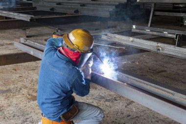 Kaynakçı, Flux Cored Arc Welding (FCAW) işlemiyle bir çelik yapı çalışması yapıyor ve endüstriyel fabrikada güvenlik için kişisel koruyucu ekipman (PPE) ile uygun şekilde giyiniyor.