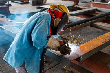 Kaynakçı, Flux Cored Arc Welding (FCAW) işlemiyle bir çelik yapı çalışması yapıyor ve endüstriyel fabrikada güvenlik için kişisel koruyucu ekipman (PPE) ile uygun şekilde giyiniyor.