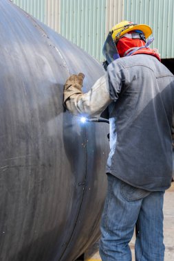 Kaynakçı, Flux Cored Arc Welding (FCAW) işlemiyle büyük bir çelik boru kaynak yapıyor ve endüstriyel fabrikada güvenlik için kişisel koruyucu ekipman (PPE) ile uygun giyiniyor..