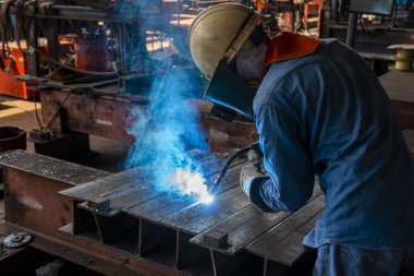 Kaynakçı, Flux Cored Arc Welding (FCAW) işlemiyle bir çelik yapı çalışması yapıyor ve endüstriyel fabrikada güvenlik için kişisel koruyucu ekipman (PPE) ile uygun şekilde giyiniyor.