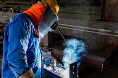 Kaynakçı, Flux Cored Arc Welding (FCAW) işlemiyle bir çelik yapı çalışması yapıyor ve endüstriyel fabrikada güvenlik için kişisel koruyucu ekipman (PPE) ile uygun şekilde giyiniyor.