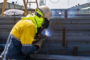 Kaynakçı, Flux Cored Arc Welding (FCAW) işlemiyle bir çelik yapı çalışması yapıyor ve endüstriyel fabrikada güvenlik için kişisel koruyucu ekipman (PPE) ile uygun şekilde giyiniyor.