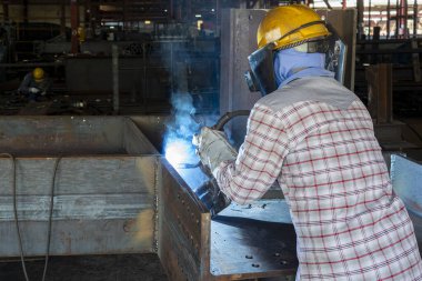 Kaynakçı, Flux Cored Arc Welding (FCAW) işlemiyle bir çelik yapı çalışması yapıyor ve endüstriyel fabrikada güvenlik için kişisel koruyucu ekipman (PPE) ile uygun şekilde giyiniyor.