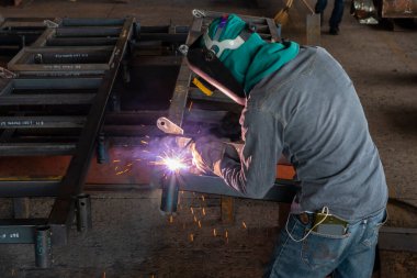 Kaynakçı, Flux Cored Arc Welding (FCAW) işlemiyle bir çelik yapı çalışması yapıyor ve endüstriyel fabrikada güvenlik için kişisel koruyucu ekipman (PPE) ile uygun şekilde giyiniyor.