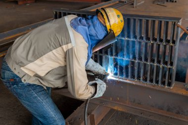 Kaynakçı, Flux Cored Arc Welding (FCAW) işlemiyle bir çelik yapı çalışması yapıyor ve endüstriyel fabrikada güvenlik için kişisel koruyucu ekipman (PPE) ile uygun şekilde giyiniyor.
