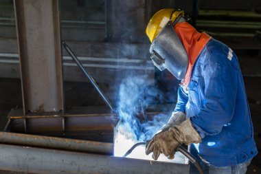 Kaynakçı, Flux Cored Arc Welding (FCAW) işlemiyle bir çelik yapı çalışması yapıyor ve endüstriyel fabrikada güvenlik için kişisel koruyucu ekipman (PPE) ile uygun şekilde giyiniyor.