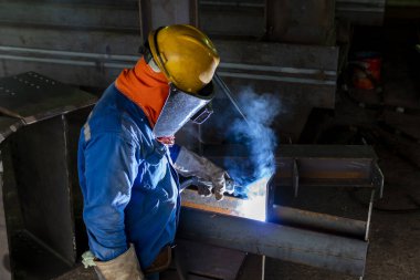 Kaynakçı, Flux Cored Arc Welding (FCAW) işlemiyle bir çelik yapı çalışması yapıyor ve endüstriyel fabrikada güvenlik için kişisel koruyucu ekipman (PPE) ile uygun şekilde giyiniyor.