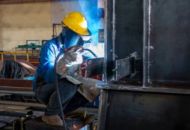 Kaynakçı, Flux Cored Arc Welding (FCAW) işlemiyle bir çelik yapı çalışması yapıyor ve endüstriyel fabrikada güvenlik için kişisel koruyucu ekipman (PPE) ile uygun şekilde giyiniyor.