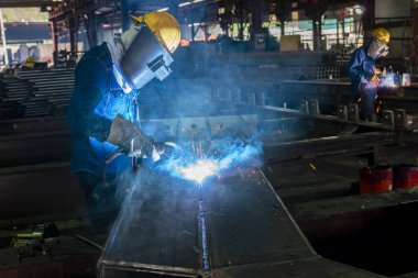 Kaynakçı, Flux Cored Arc Welding (FCAW) işlemiyle bir çelik yapı çalışması yapıyor ve endüstriyel fabrikada güvenlik için kişisel koruyucu ekipman (PPE) ile uygun şekilde giyiniyor.